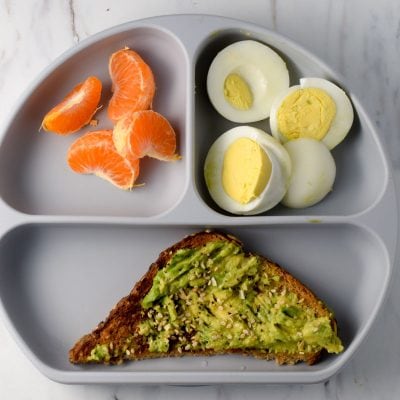 healthy breakfast for toddlers shown on a grey plate avocado toast, hard boiled eggs, and oranges